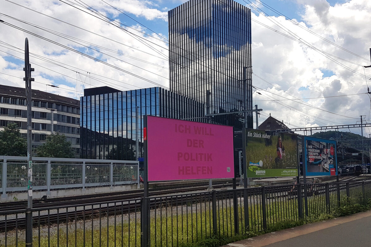 Auf einer der Werbeflächen am Bahnhof in St. Gallen ist ein Plakat des Projekts ¡ausrufezeichen! zu sehen. Auf dem Plakat steht: Ich will der Politik helfen. Im Hintergrund ist das St. Galler Rathaus zu sehen.