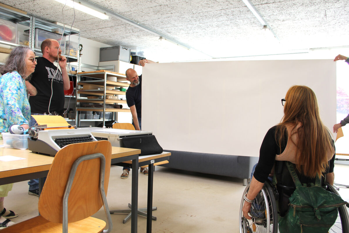 Eine Person im Rollstuhl schaut auf ein Papier im Format F12, dass zwei Personen hochhalten.