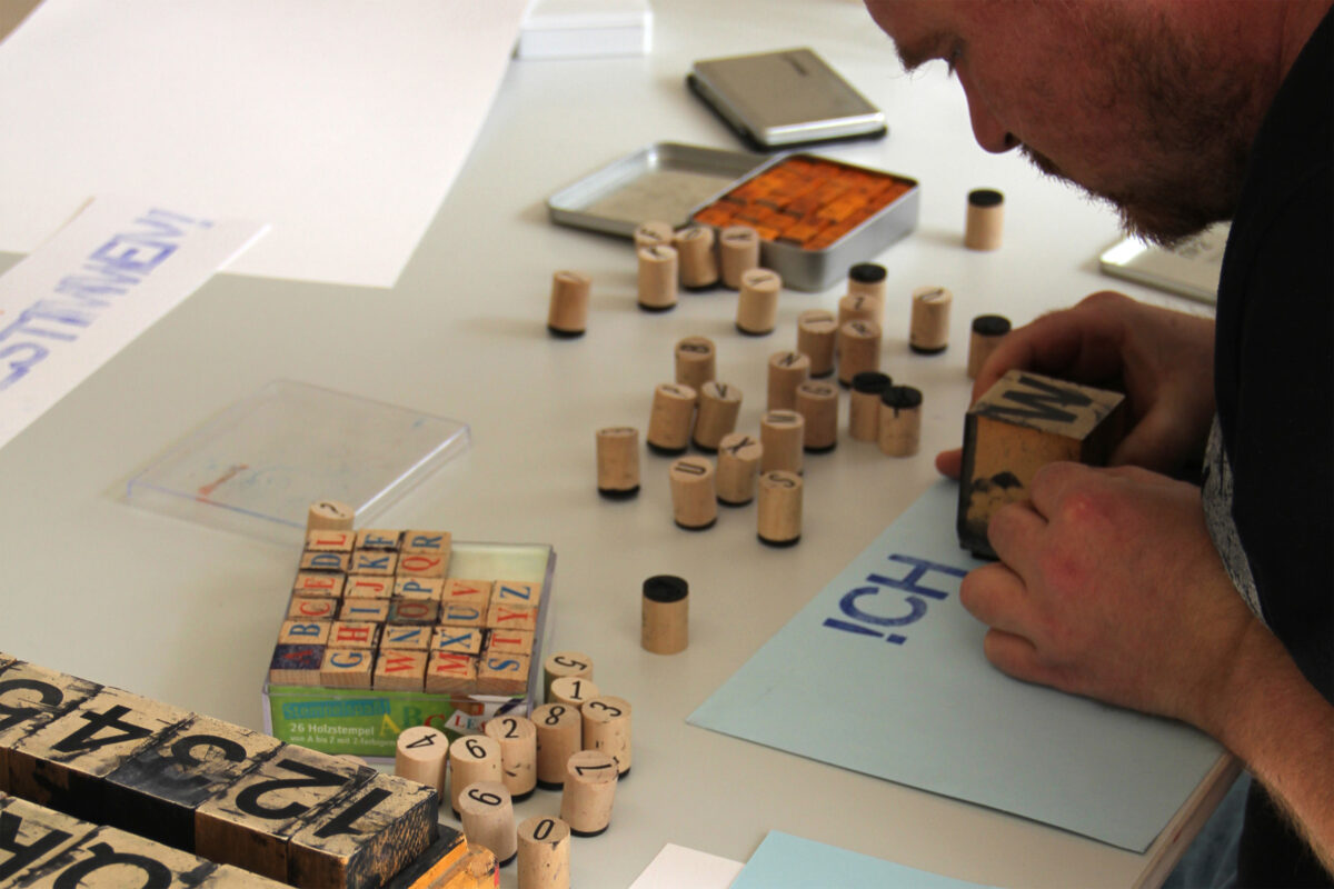 Eine Person schreibt den Text ihres Plakats mit verschieden grossen Stempeln auf ein farbiges Blatt Papier.